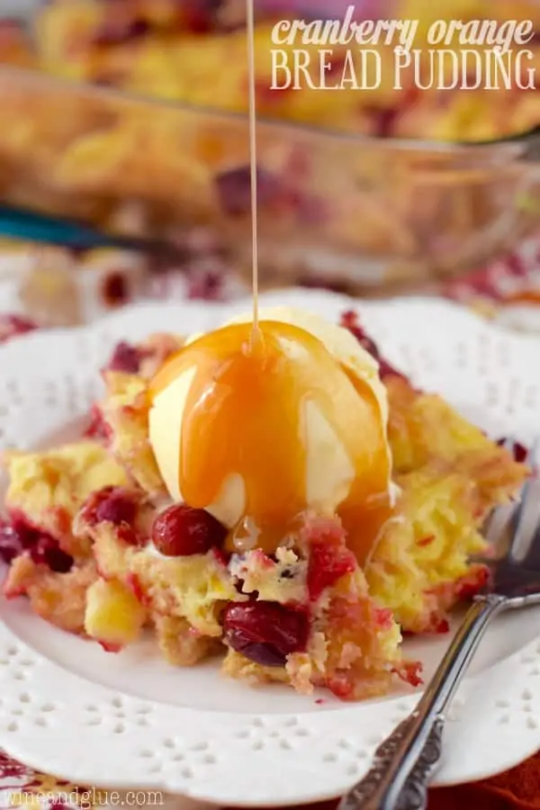 On a white plate, the Cranberry Orange Bread Pudding is topped with a scoop of vanilla ice cream and caramel is being drizzled right on top. 