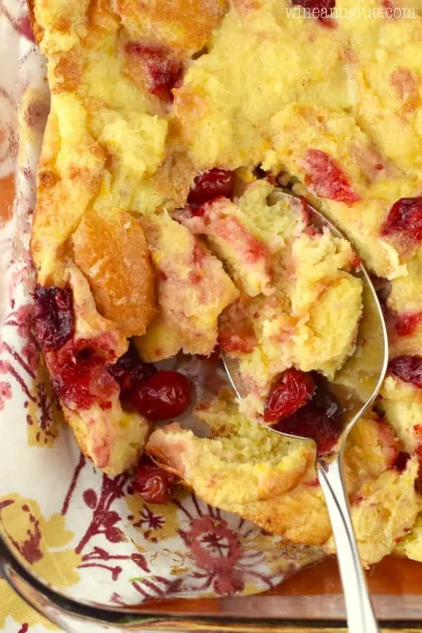 In a glass casserole dish, a spoon is scooping into the Cranberry Orange Bread Pudding showing the speckles of red from the cranberries. 
