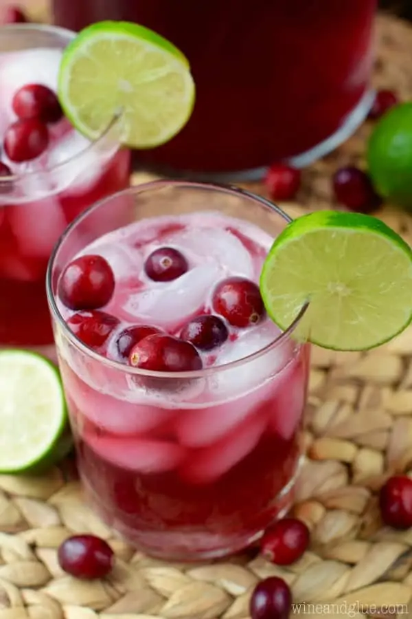Two glasses of the Cranberry Beergaritas with a dark maroon color with ice cubes, whole cranberries, and a slice of lime on the rim. 