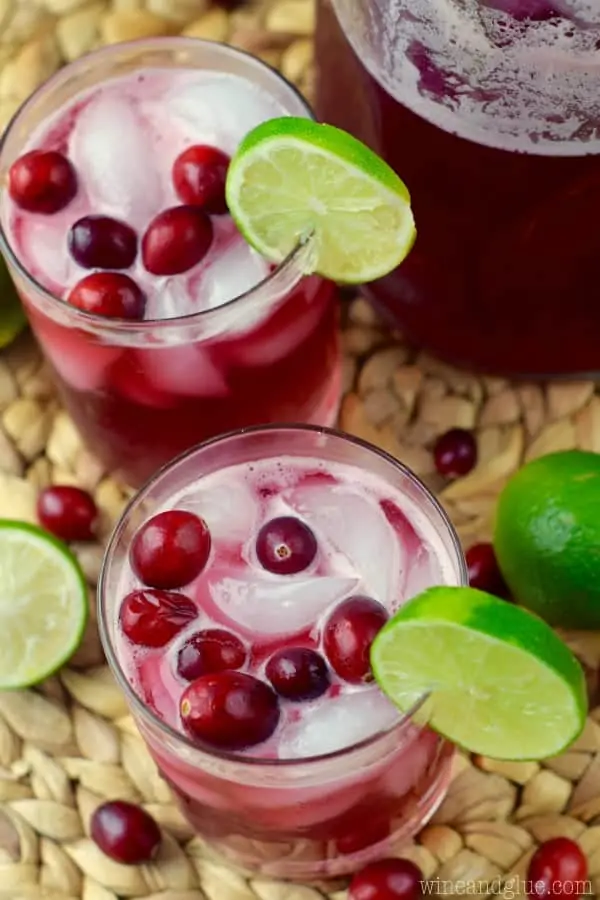 Two glasses of the Cranberry Beergaritas with a dark maroon color with ice cubes, whole cranberries, and a slice of lime on the rim. 