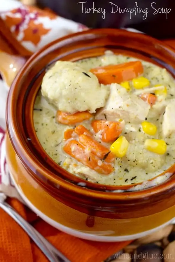 In a brown ceramic bowl, the Turkey Dumpling Soup has chunks of turkey, balls of dumpling, slices of carrots, and corn in a creamy broth. 