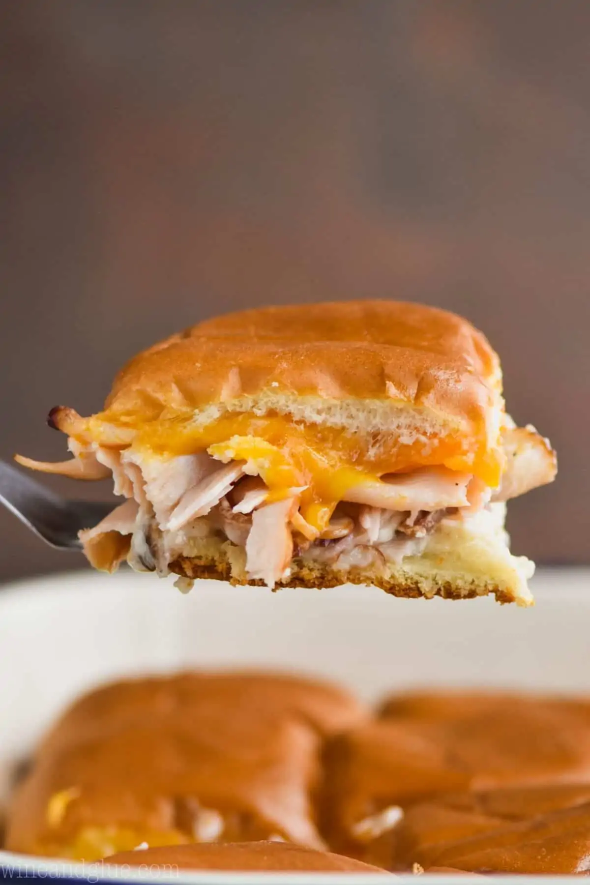 a bacon ranch turkey slider being pulled out of a baking dish of other sliders with melted cheddar cheese and turkey visible - 16