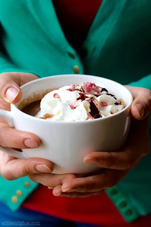 A woman holding a big mug full of Peppermint Hot Chocolate topped with whipped cream, crushed peppermint, and drizzled with chocolate. 