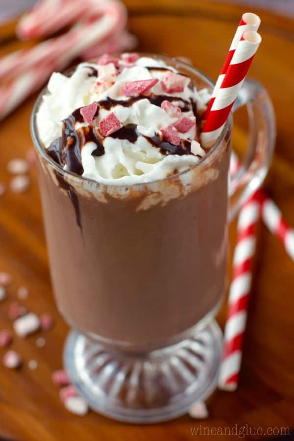 In a glass mug, the Peppermint Hot Chocolate is topped with whipped cream, crushed peppermint, and chocolate syrup with two paper straws.