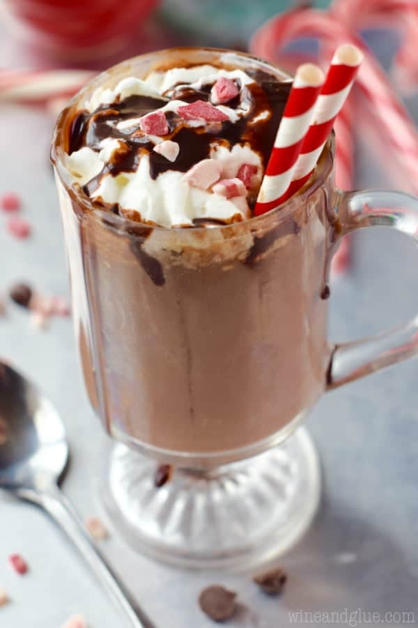 In a glass mug, the Peppermint Hot Chocolate is topped with whipped cream, crushed peppermint, and chocolate syrup with two paper straws.