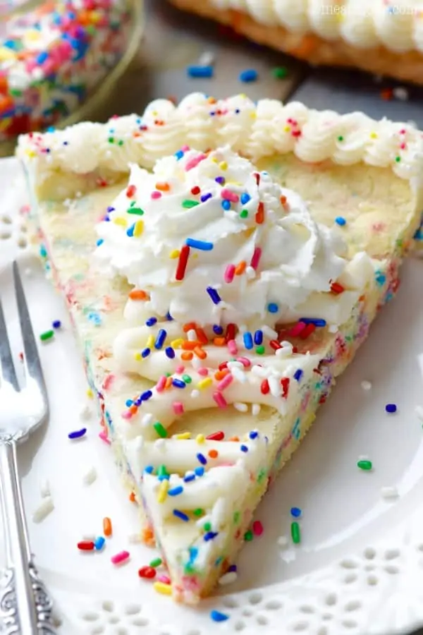 On a white plate, the Funfetti Cookie Cake has white frosting on the edge of the cookie cake with a star tip and whipped cream in the middle topped with rainbow sprinkles. 