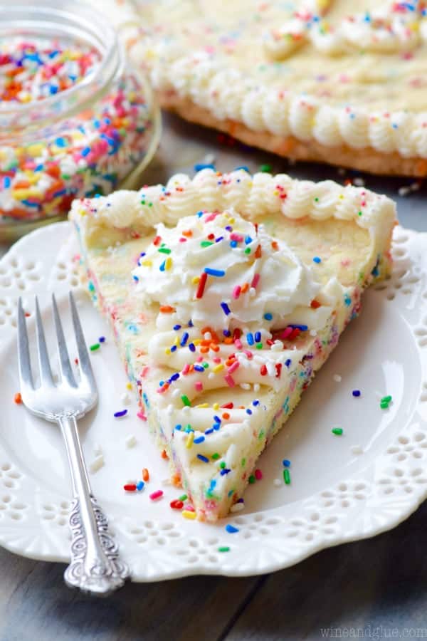 On a white plate, the Funfetti Cookie Cake has white frosting on the edge of the cookie cake with a star tip and whipped cream in the middle topped with rainbow sprinkles. 
