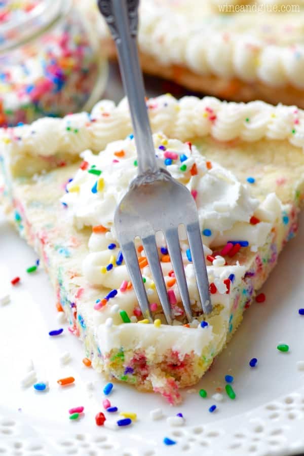 A fork digging into the Funfetti Cookie Cake has white frosting on the edge of the cookie cake with a star tip and whipped cream in the middle topped with rainbow sprinkles. 