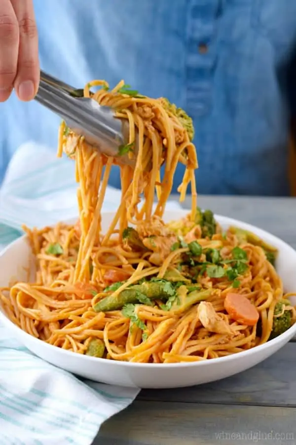 In a bowl, the Sriracha Noodles are being picked up with a pair of metal tongs. 