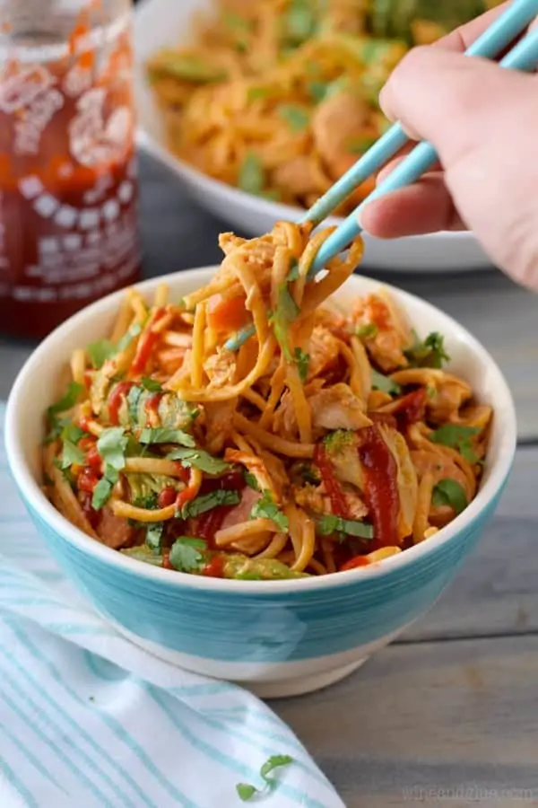 A pair of blue chopsticks is picking up some of the Sriracha Noddles from a blue bowl. 