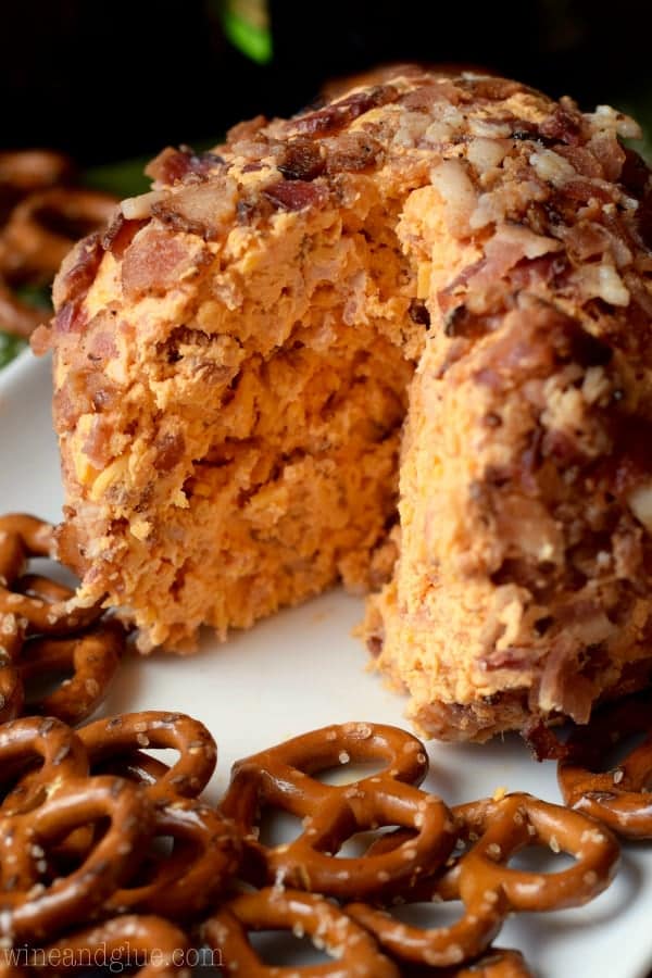 A large slice of the Buffalo Bacon Cheeseball shows the buffalo red color inside, has a crust of minced bacon, and served with pretzels. 