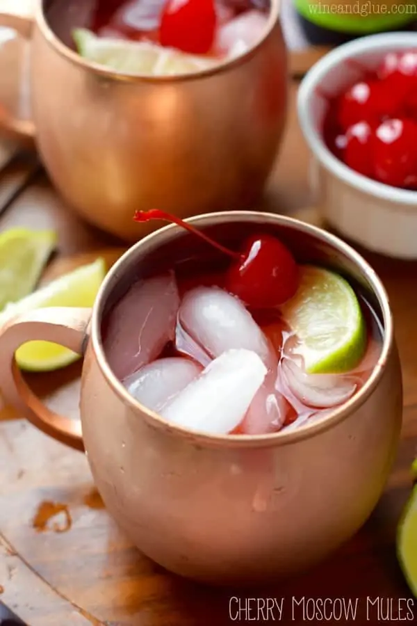 In copper mugs, the Cherry Moscow Mules has cubed ice, red tint, a maraschino cherry, and a sliced lime. 