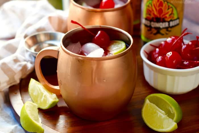In copper mugs, the Cherry Moscow Mules has cubed ice, red tint, a maraschino cherry, and a sliced lime. 