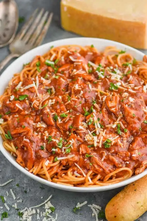 a large white serving bowl with chicken spaghetti, garnished with parsley and parmesan cheese with a block of fresh parmesan in the background