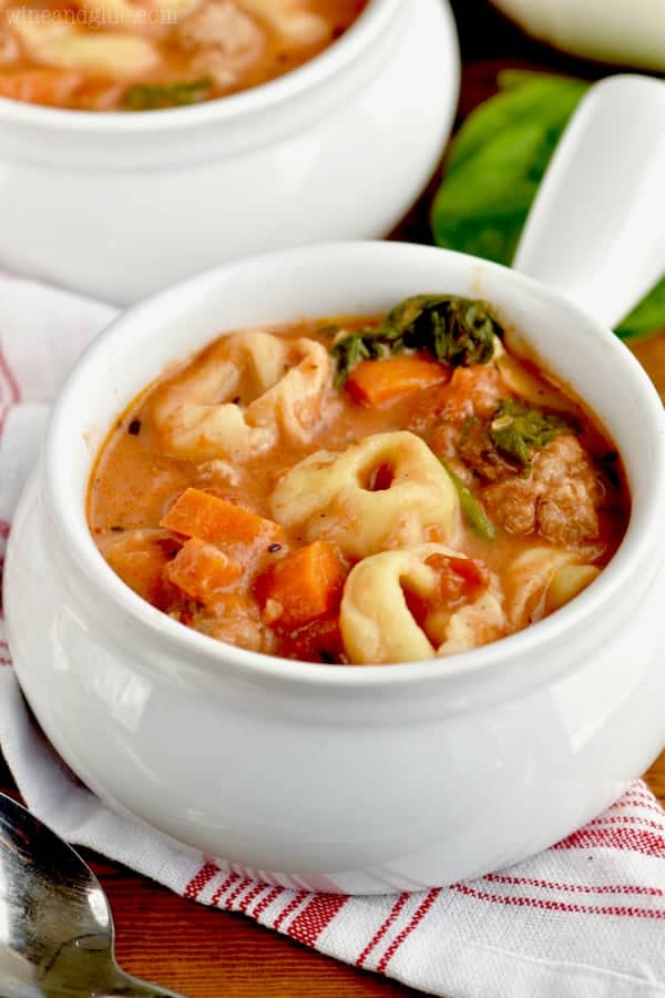 In a small white bowl, the Slow Cooker Creamy Tortellini Soup has spinach, sausage, and chunks of carrots in a creamy tomato soup. 