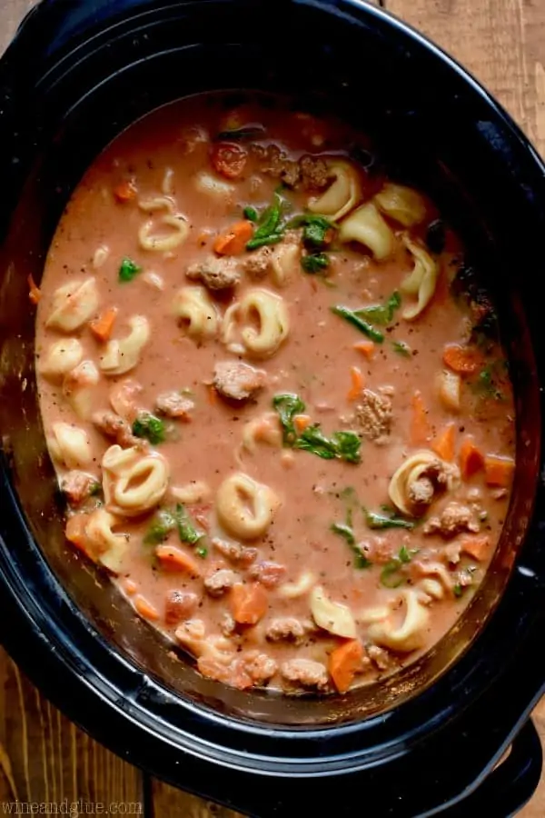 An overhead photo of a crockpot full of the Slow Cooker Creamy Tortellini Soup with Tortellini, spinach, chunks of sausage, and large cuts of carrots in a creamy tomato broth. 