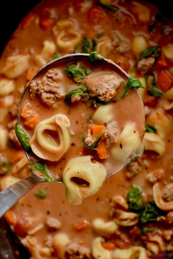 An overhead photo of the ladle scooping some of the Slow Cooker Creamy Tortellini Soup showing the chunks of sausage, cooked spinach, tortellini, and large carrots. 