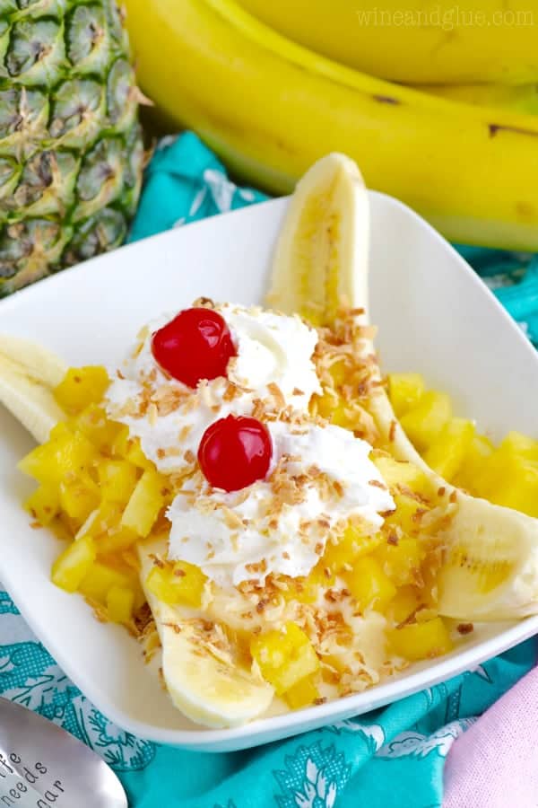 In a white dish, the Tropical Banana Split has chunks of pineapple, a banana sliced in half, two whipped cream dollops, maraschino cherries, and toasted coconut flakes. 