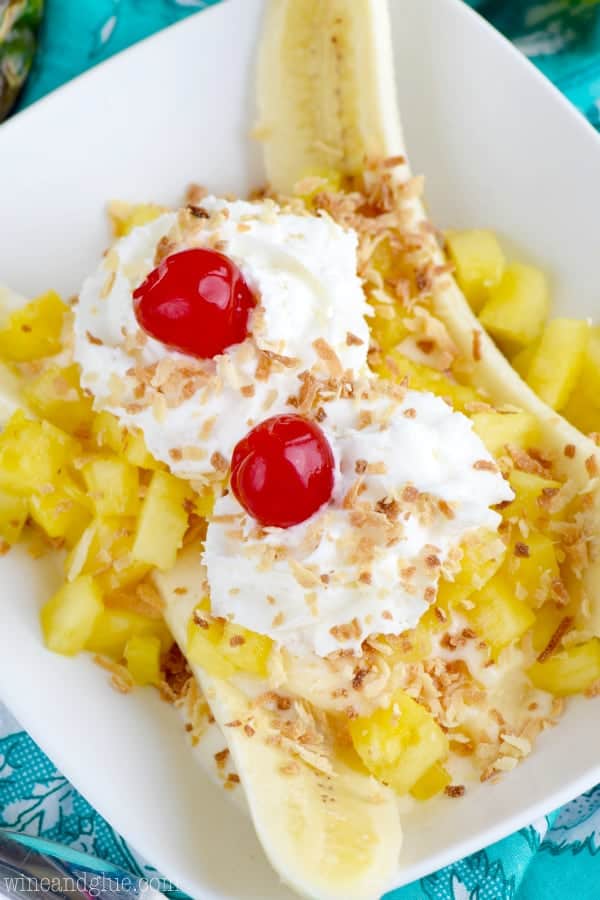 In a white dish, the Tropical Banana Split has chunks of pineapple, a banana sliced in half, two whipped cream dollops, maraschino cherries, and toasted coconut flakes. 