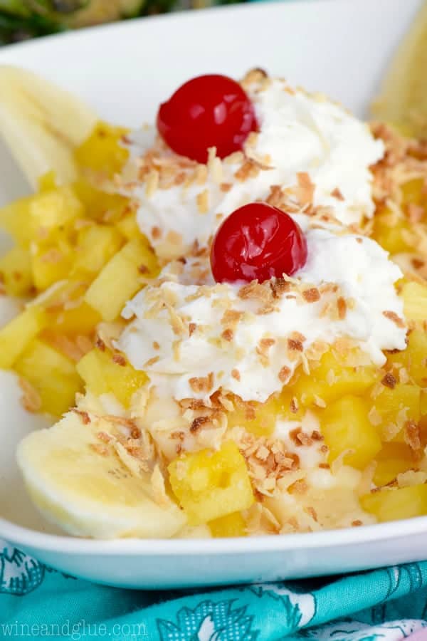 In a white dish, the Tropical Banana Split has chunks of pineapple, a banana sliced in half, two whipped cream dollops, maraschino cherries, and toasted coconut flakes. 