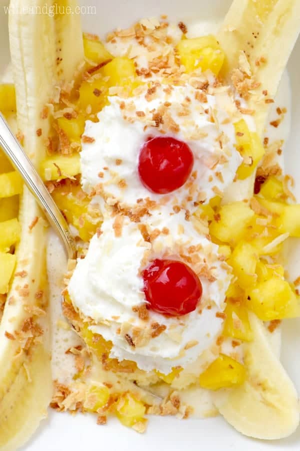 In a white dish, the Tropical Banana Split has chunks of pineapple, a banana sliced in half, two whipped cream dollops, maraschino cherries, and toasted coconut flakes. 