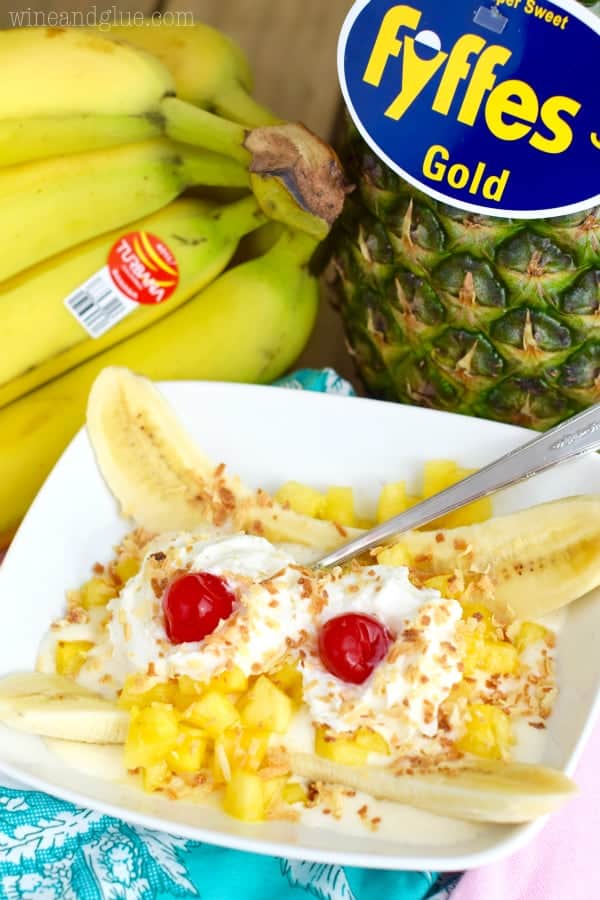 In a white dish, the Tropical Banana Split has chunks of pineapple, a banana sliced in half, two whipped cream dollops, maraschino cherries, and toasted coconut flakes. 
