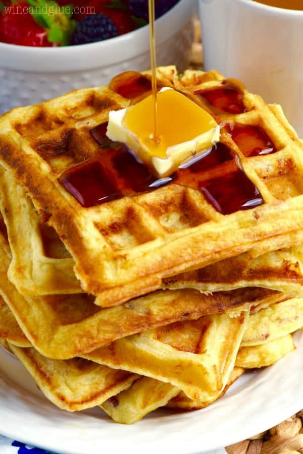 A stack of Bacon Cheddar Sour Cream Waffle has a a slice of butter and syrup being poured on top. 