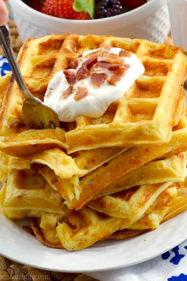 A fork digging into the Bacon Cheddar Sour Cream Waffles which has a golden brown color and topped with a dollop of sour cream and minced bacon. 