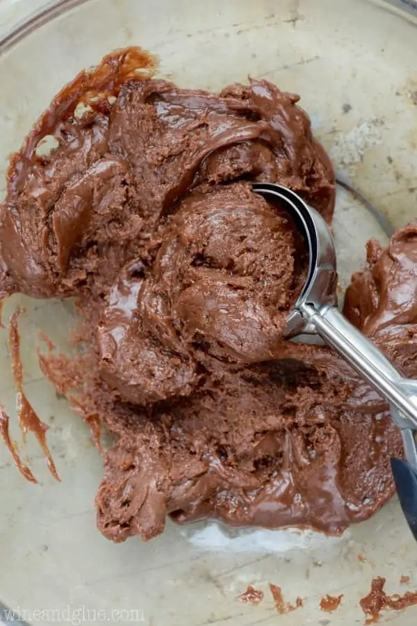 In a glass bowl, the creamy chocolate fudge is being scooped with an ice cream scooper.