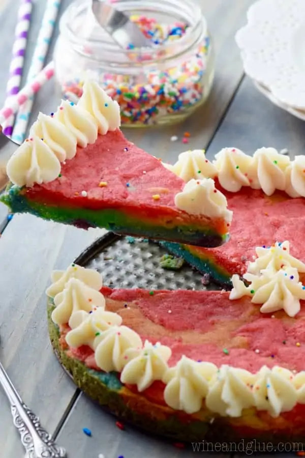 A slice of the Rainbow Sugar Cookie Cake is being taken out of the cake showing the distinct layers of red, orange, yellow, green, blue, and purple (in that order from top to bottom). 