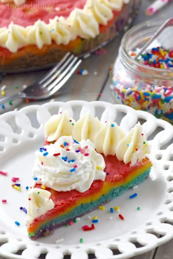 A slice of the Rainbow Sugar Cookie cake is on a white plate showing the colors of the rainbow and topped with white frosting on the edges and rainbow sprinkles. 