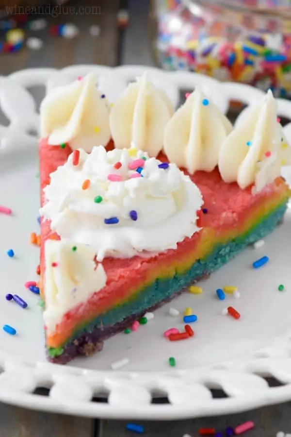 A slice of the Rainbow Sugar Cookie cake is on a white plate showing the colors of the rainbow and topped with white frosting on the edges and rainbow sprinkles. 