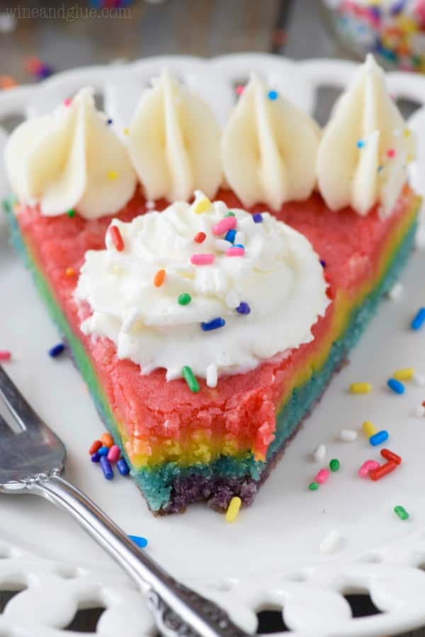A slice of the Rainbow Sugar Cookie Cake is topped with white frosting on the edge with a star tip, a dollop of whipped cream in the middle, and rainbow sprinkles. 