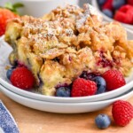 Pinterest graphic for Berry French Toast Casserole recipe. Image is photo of a piece of Berry French Toast Casserole served on a plate garnished with berries. Bowl of berries and cups of coffee on counter beside plate. Text says, "super easy Berry French Toast Casserole simplejoy.com"