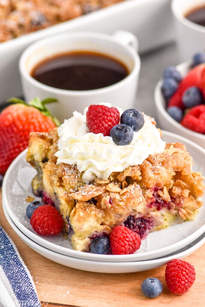 Overhead photo of a piece of Berry French Toast Casserole served on a plate garnished with berries and whipped cream. Bowl of berries and cup of coffee on counter.