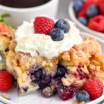 Close up photo of a piece of Berry French Toast Casserole served on a plate garnished with berries and whipped cream. Cup of coffee and bowl of berries behind plate.