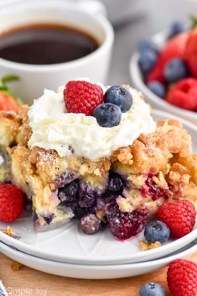 Close up photo of a piece of Berry French Toast Casserole served on a plate garnished with berries and whipped cream. Cup of coffee and bowl of berries behind plate.