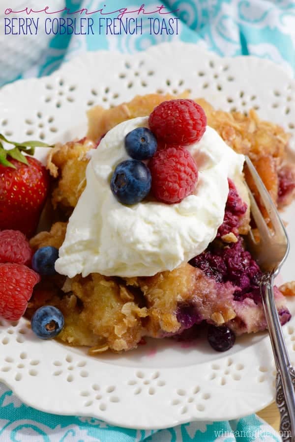On a white plate, the Overnight Berry Cobble French Toast is topped with a dollop of freshly made whipped cream and surrounded by blueberries, strawberries, and raspberries. 