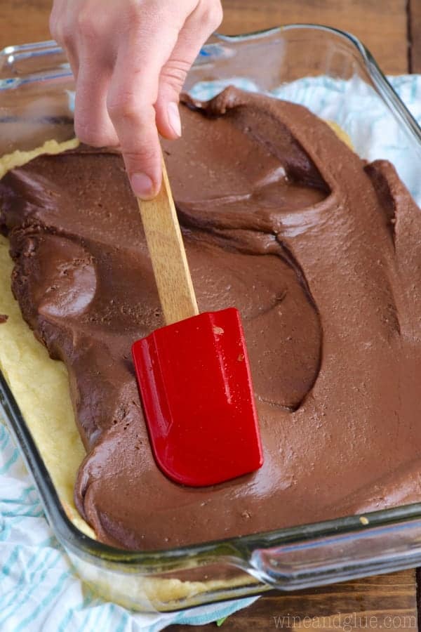 In a glass casserole dish, a layer of baked sugar cookies is being topped with some fudge that is being spread by a spatula. 