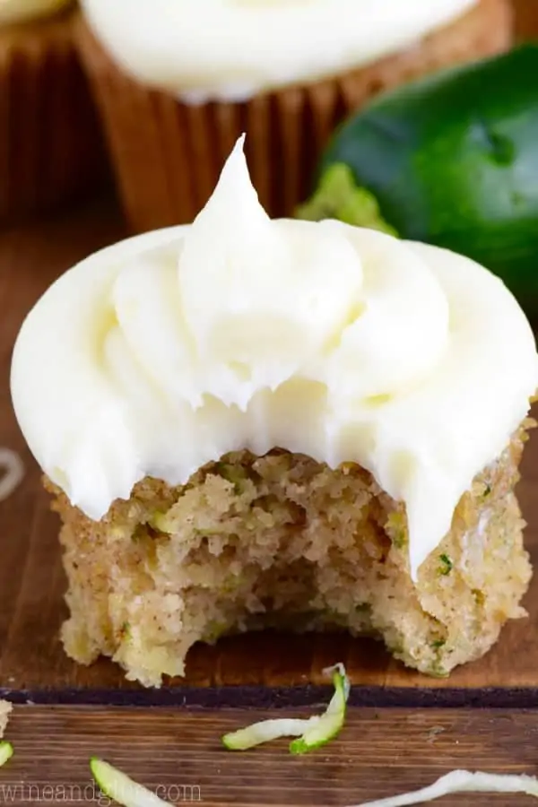 In a cupcake tin, the Zucchini Cupcake has a large bite in it showing the a dark golden brown color and topped with a fluffy cream cheese frosting. 