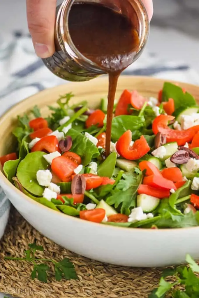 a hand pouring balsamic salad dressing onto a salad with mixed greens, tomatoes, red peppers, feta cheese, and kalamata olives