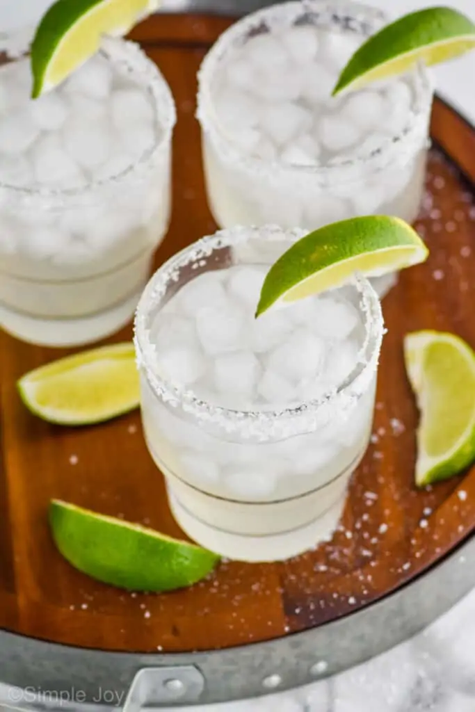 overhead view of three tumblers with margarita recipe in them, ice, salted rims, lime wedges on a wooden tray that has salt around it and three lime wedges