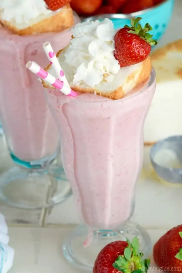 In a milkshake glass, the pink Boozy Strawberry Shortcake Milkshake is topped with a small slice of a strawberry shortcake, a sliced strawberry, and whipped cream.