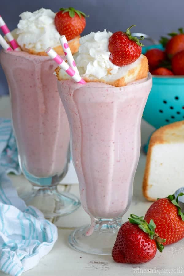 In a milkshake glass, the pink Boozy Strawberry Shortcake Milkshake is topped with a small slice of a strawberry shortcake, a sliced strawberry, and whipped cream.