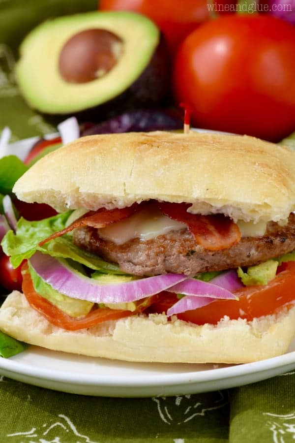 The California Club Burger is in between two ciabatta buns with turkey bacon, turkey burger, lettuce, tomatoes, avocado, and tomatoes with a side of salad. 
