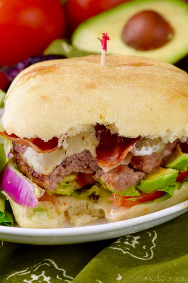 The California Club Burger is in between two ciabatta buns with turkey bacon, turkey burger, lettuce, tomatoes, avocado, and tomatoes with a side of salad. 