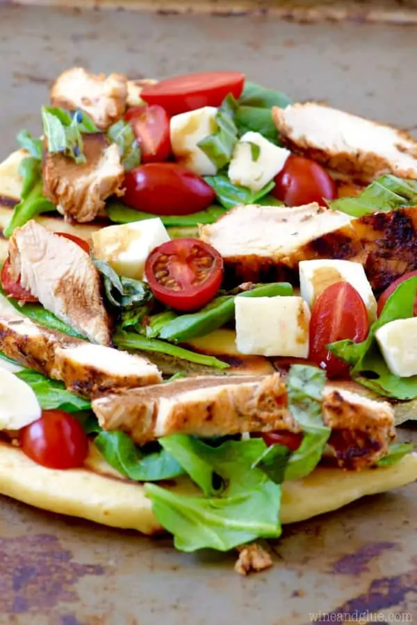 On a baking sheet, the Grilled Chicken Capese Flatbread has sliced cherry tomatoes, basil, grilled cheese, and mozzarella. 