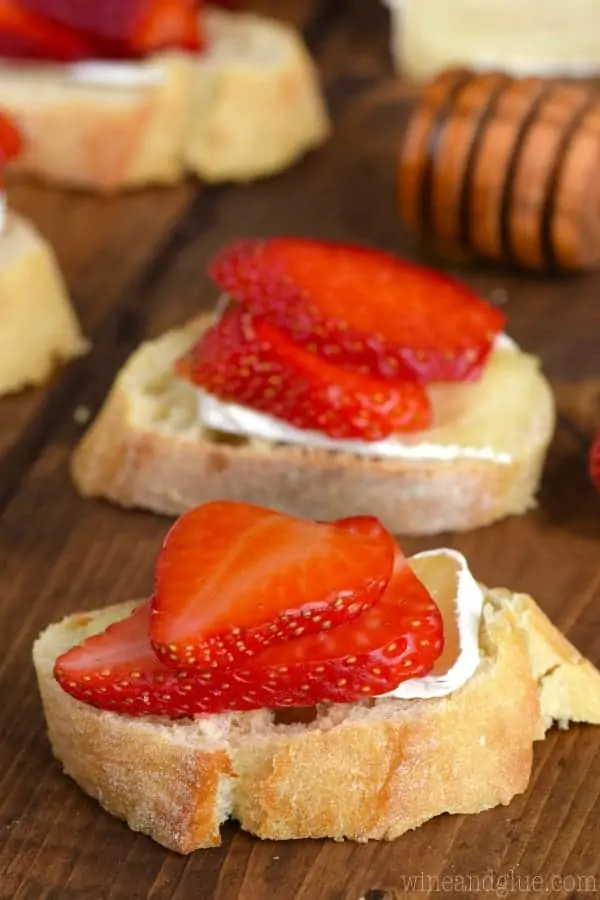 On a slice of baguette, a slice of Brie is topped with two thin slices of strawberry and honey is drizzled on top to make the Honey, Strawberry, and Brie Bruschetta. 