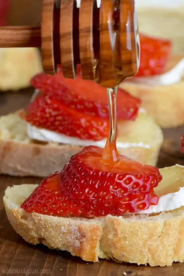 On a slice of baguette, a slice of Brie is topped with two thin slices of strawberry and honey is drizzled on top to make the Honey, Strawberry, and Brie Bruschetta. 