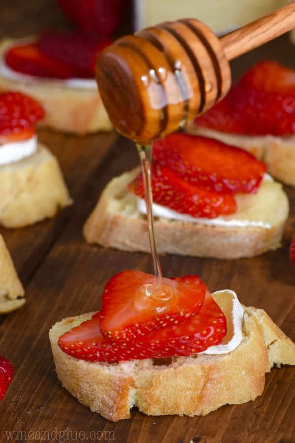 On a slice of baguette, a slice of Brie is topped with two thin slices of strawberry and honey is drizzled on top to make the Honey, Strawberry, and Brie Bruschetta. 